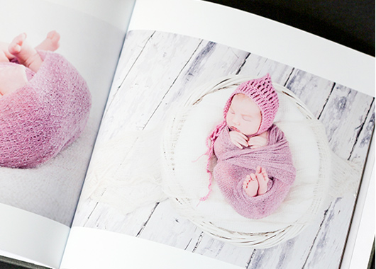 Square baby photo book open to pages featuring photos of a child wrapped in pink blanket.
